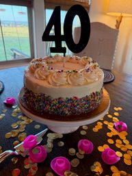 Vanilla frosted birthday cake with a black '40' topper, piped rosettes and colorful sprinkles on a white pedestal, surrounded by gold confetti and pink toy cowboy-hat party favors on a wooden dining table by a window with a grassy yard view.