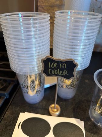 Stacked clear plastic party cups on a kitchen counter with a small chalkboard sign reading 'Ice in cooler' and round black stickers nearby