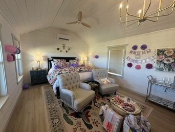 Bright cozy farmhouse-style bedroom with white shiplap walls, floral bedding on a dark wood bed, tufted armchairs, patterned rug, ceiling fan, side tables and birthday decorations.