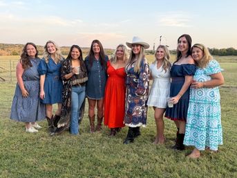 Nine women in colorful summer and western-style dresses, boots and a cowboy hat posing together on a grassy rural field at sunset, smiling and holding drinks.