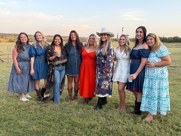 Nine women in colorful summer and western-style dresses, boots and a cowboy hat posing together on a grassy rural field at sunset, smiling and holding drinks.