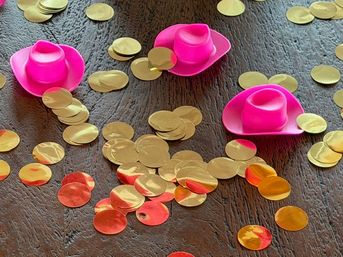 Playful party scene: three mini bright pink cowboy hat favors scattered among metallic gold and rose‑gold circular confetti on a dark wooden table.