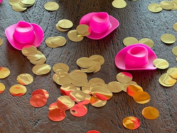 Playful party scene: three mini bright pink cowboy hat favors scattered among metallic gold and rose‑gold circular confetti on a dark wooden table.
