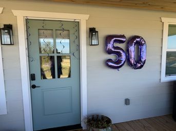 Home front porch with a light-blue door, purple hanging streamers and metallic purple “50” balloons on gray siding above a wooden deck for a 50th birthday celebration