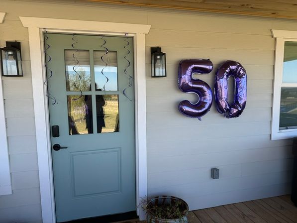 Home front porch with a light-blue door, purple hanging streamers and metallic purple “50” balloons on gray siding above a wooden deck for a 50th birthday celebration