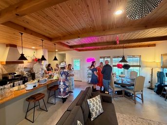 Cozy open-plan home interior with wood-beamed ceiling and pendant lights, guests mingling around a kitchen island and decorated dining table under a pink neon sign.