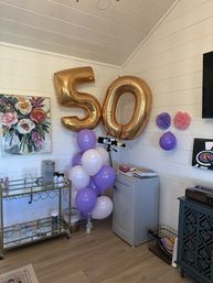 Cozy shiplap room decorated for a 50th celebration with large gold '50' foil balloons, a purple and white balloon cluster, mini fridge, bar cart with glasses, floral wall art and paper pom-poms.