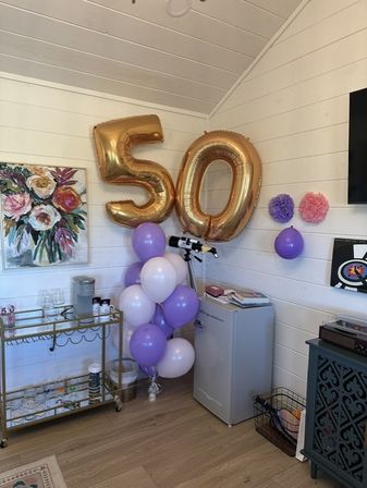 Cozy shiplap room decorated for a 50th celebration with large gold '50' foil balloons, a purple and white balloon cluster, mini fridge, bar cart with glasses, floral wall art and paper pom-poms.