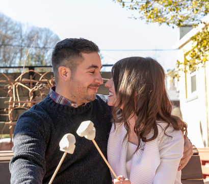Smiling couple cuddled on an outdoor bench, sharing a cozy red blanket and roasting marshmallows on skewers on a sunny day.