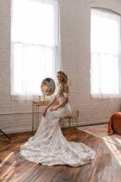 Woman in a lace off-the-shoulder wedding gown with long train seated at a gold vanity mirror in a sunlit white-brick loft with tall windows and hardwood floors