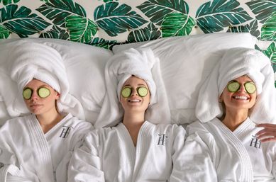 Three friends in white spa robes with towel turbans and cucumber slices over their eyes, smiling and relaxing on a bed against tropical green leaf wallpaper — spa day self-care.