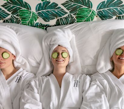 Three friends in white spa robes with towel turbans and cucumber slices over their eyes, smiling and relaxing on a bed against tropical green leaf wallpaper — spa day self-care.