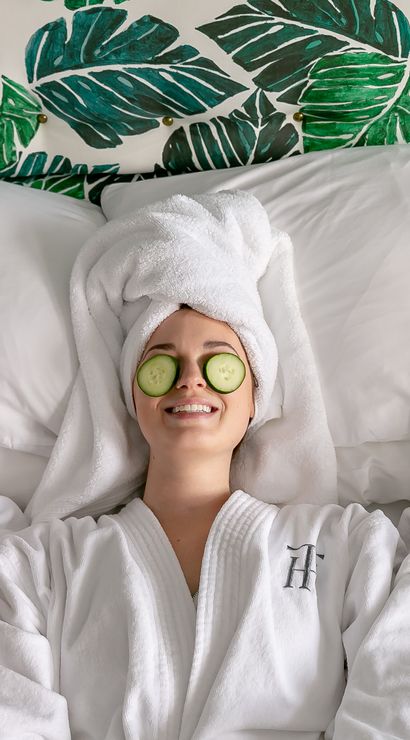 Three friends in white spa robes with towel turbans and cucumber slices over their eyes, smiling and relaxing on a bed against tropical green leaf wallpaper — spa day self-care.