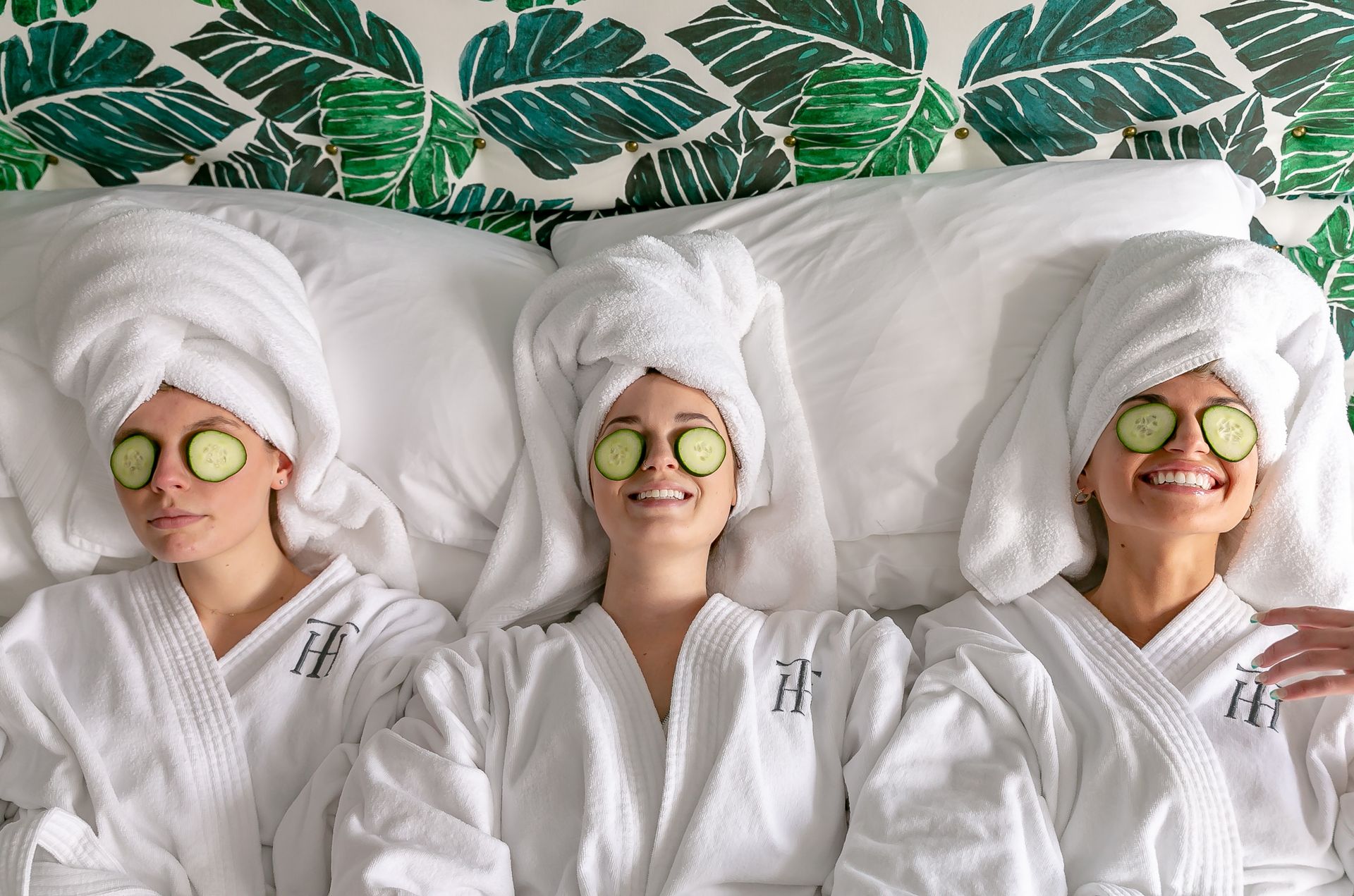 Three friends in white spa robes with towel turbans and cucumber slices over their eyes, smiling and relaxing on a bed against tropical green leaf wallpaper — spa day self-care.