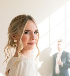 Sunlit close-up of a smiling bride with a blonde updo and statement earrings, soft indoor wedding portrait with a blurred groom in a suit by a window