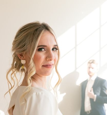 Sunlit close-up of a smiling bride with a blonde updo and statement earrings, soft indoor wedding portrait with a blurred groom in a suit by a window