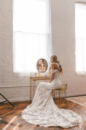 Bride in a lace off-the-shoulder wedding gown seated at a gold vanity in a sunlit white-brick loft, gazing into a round mirror.