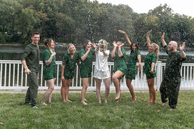 Lakeside celebration on a white deck — friends in matching green outfits and one in white pop a champagne bottle, spraying bubbly and cheering in front of a wooded lake.