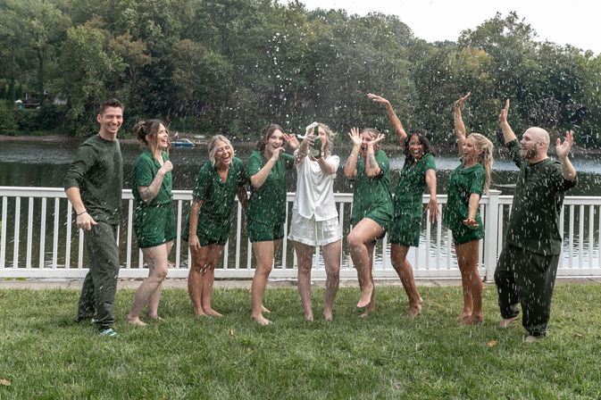 Lakeside celebration on a white deck — friends in matching green outfits and one in white pop a champagne bottle, spraying bubbly and cheering in front of a wooded lake.
