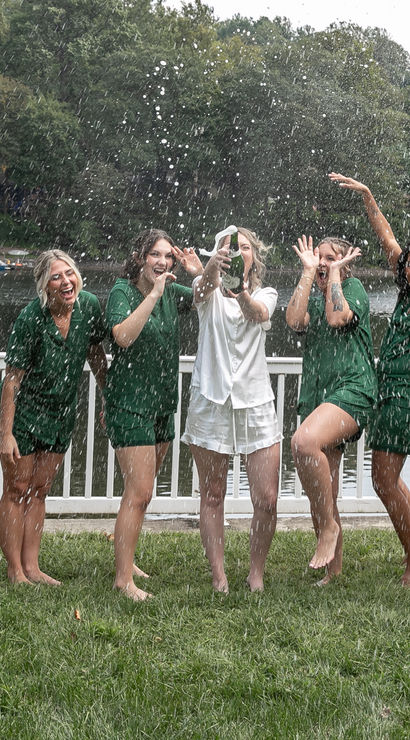 Lakeside celebration on a white deck — friends in matching green outfits and one in white pop a champagne bottle, spraying bubbly and cheering in front of a wooded lake.