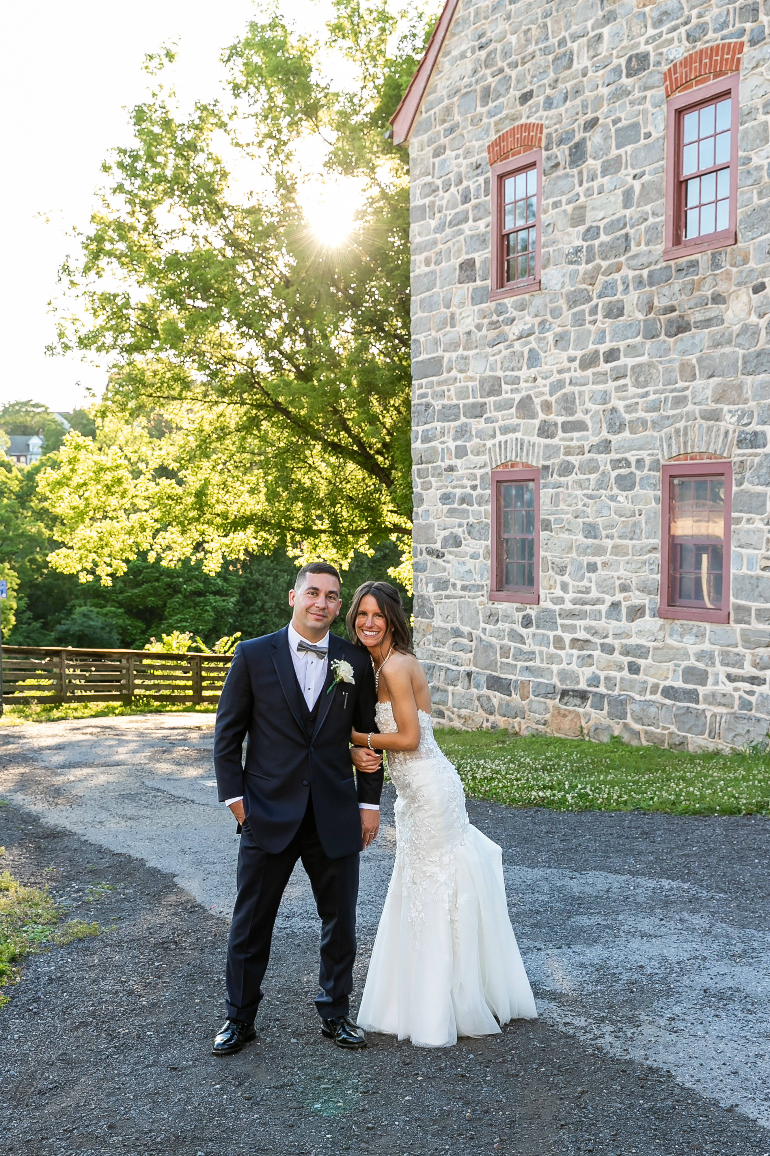 Smiling bride and groom pose beside a rustic stone building with golden sun flare and green trees, outdoor wedding portrait.