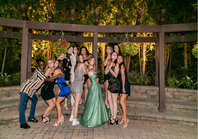 Group of teens in formal dresses and a suit posing playfully under a lit wooden pergola with hanging flower baskets in an outdoor evening garden, prom/formal event photo