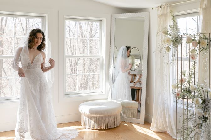 Veiled bride in a lace wedding gown standing by large windows in a sunlit bridal suite, reflected in a full‑length mirror beside a tufted ottoman and floral display.
