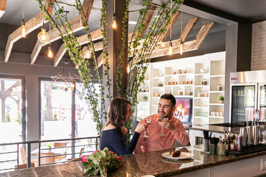 Couple toasting cocktails at a marble bar in a modern rustic-chic restaurant with ivy-wrapped beams, hanging Edison bulbs and a dessert on the counter
