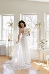 Sunlit bridal portrait in a bright room — bride in lace wedding gown and veil holding a white rose bouquet by tall windows with blush-and-white floral stands.