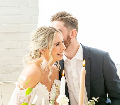 Bride in lace off-shoulder gown laughing as groom whispers at a candlelit wedding table with blush roses, eucalyptus and soft florals against a white brick loft wall
