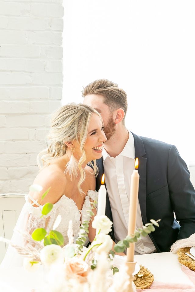 Bride in lace off-shoulder gown laughing as groom whispers at a candlelit wedding table with blush roses, eucalyptus and soft florals against a white brick loft wall