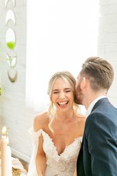 Joyful wedding photo of a bride laughing as the groom whispers in her ear, bride in a floral lace off-shoulder gown and groom in a navy suit in a bright indoor venue.