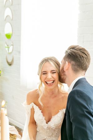 Joyful wedding photo of a bride laughing as the groom whispers in her ear, bride in a floral lace off-shoulder gown and groom in a navy suit in a bright indoor venue.