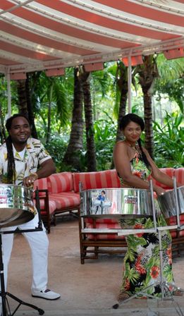 Two musicians playing steelpan drums under a red-and-white striped canopy in a lush tropical outdoor lounge with palm trees and red-striped cushioned seating