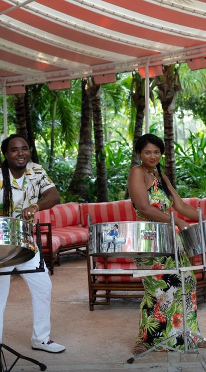 Two musicians playing steelpan drums under a red-and-white striped canopy in a lush tropical outdoor lounge with palm trees and red-striped cushioned seating