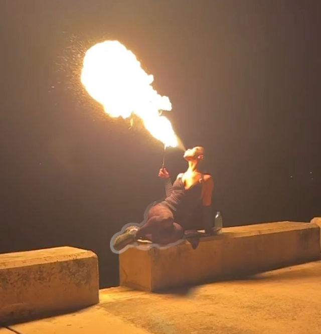 Fire-breather exhaling a huge fireball at night while reclining on a concrete pier or seawall, the orange flame lighting the dark waterfront with a bottle nearby.