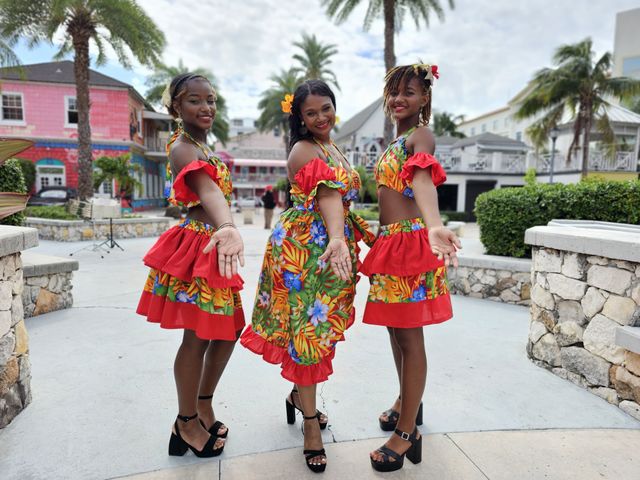 Three dancers in vibrant red-and-floral Caribbean costumes and high heels pose with outstretched hands in a palm-lined, pastel-colored waterfront plaza.