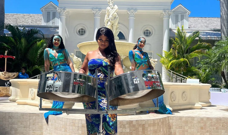 Three women in mermaid-style outfits play steelpan drums poolside in front of a grand white villa fountain and palm trees at a sunny tropical resort.