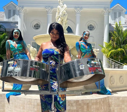 Three women in mermaid-style outfits play steelpan drums poolside in front of a grand white villa fountain and palm trees at a sunny tropical resort.