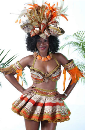 Smiling woman in a vibrant Caribbean-style carnival costume with an oversized orange-and-gold feathered headdress, beaded bra top, matching flared skirt and feathered arm cuffs, tropical palms in the background.