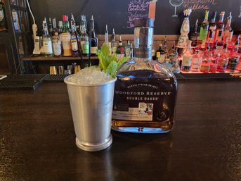 Silver julep cup piled with crushed ice and a mint sprig next to a Woodford Reserve Double Oaked bourbon bottle on a dark wood bar counter, with liquor bottles and a chalkboard menu in the background.