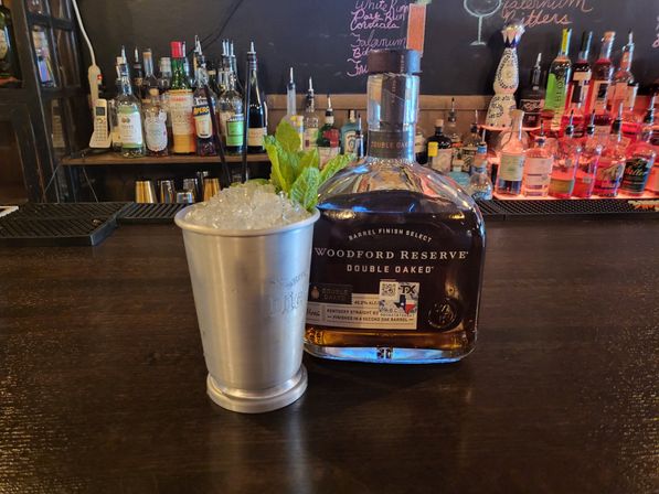 Silver julep cup piled with crushed ice and a mint sprig next to a Woodford Reserve Double Oaked bourbon bottle on a dark wood bar counter, with liquor bottles and a chalkboard menu in the background.