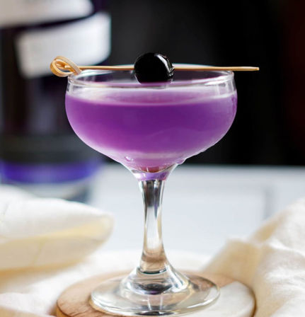 Vibrant purple cocktail in a coupe glass topped with a dark cherry on a wooden pick, resting on a coaster with soft white linen and a blurred bar background.