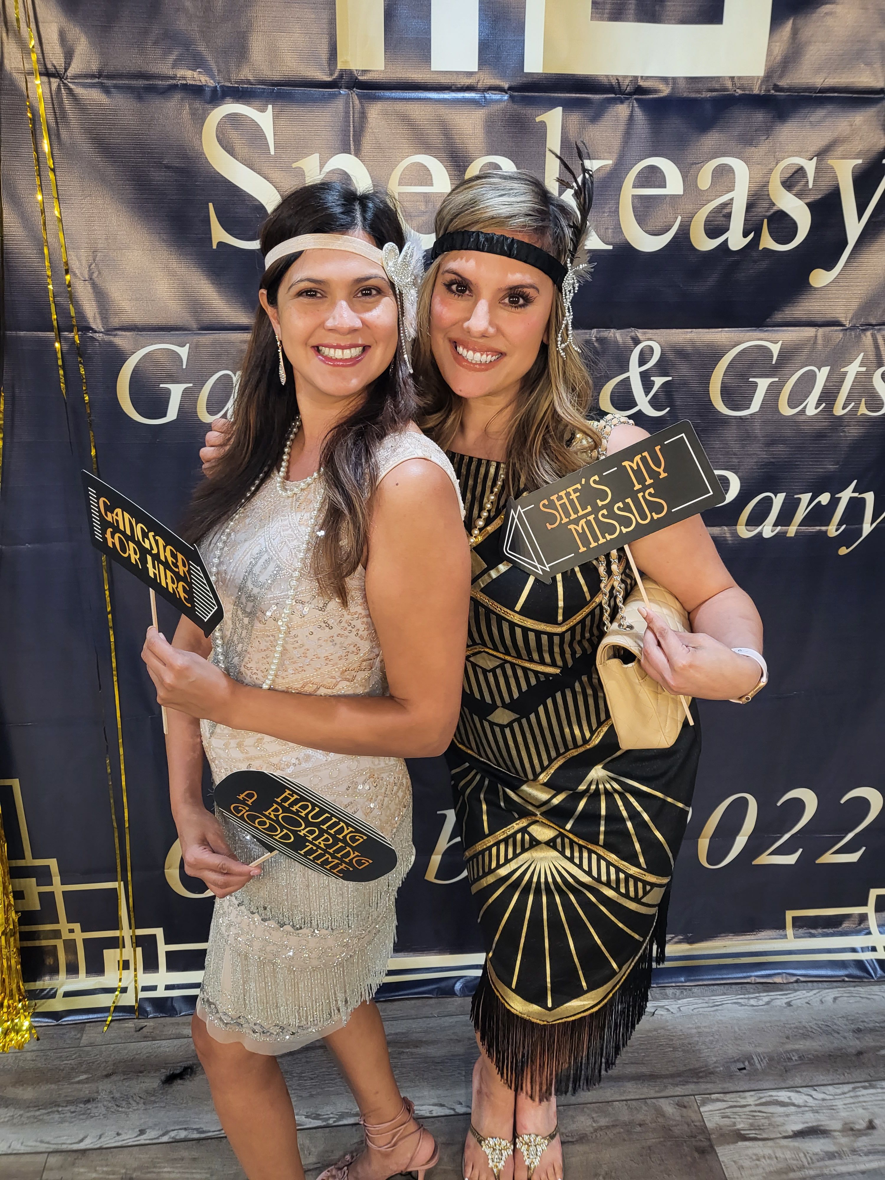 Two smiling women in 1920s flapper dresses and headbands pose with photo‑booth signs at a Gatsby speakeasy party backdrop.