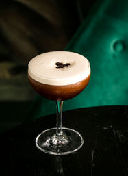 Espresso martini in a coupe glass with creamy foam and three coffee beans on top, set on a black table with a green velvet background.