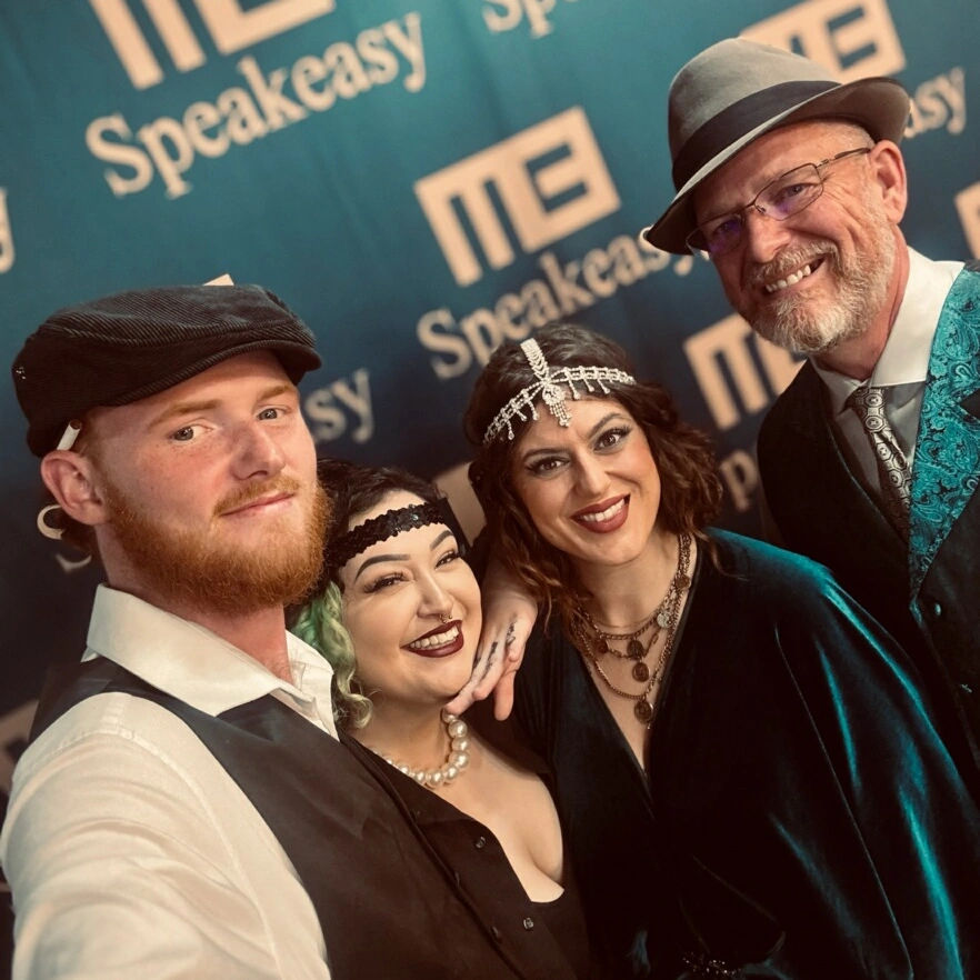 Four people in 1920s speakeasy-style attire—men in hats and vests, women in jeweled headbands and pearls—smiling at a retro event photo backdrop.