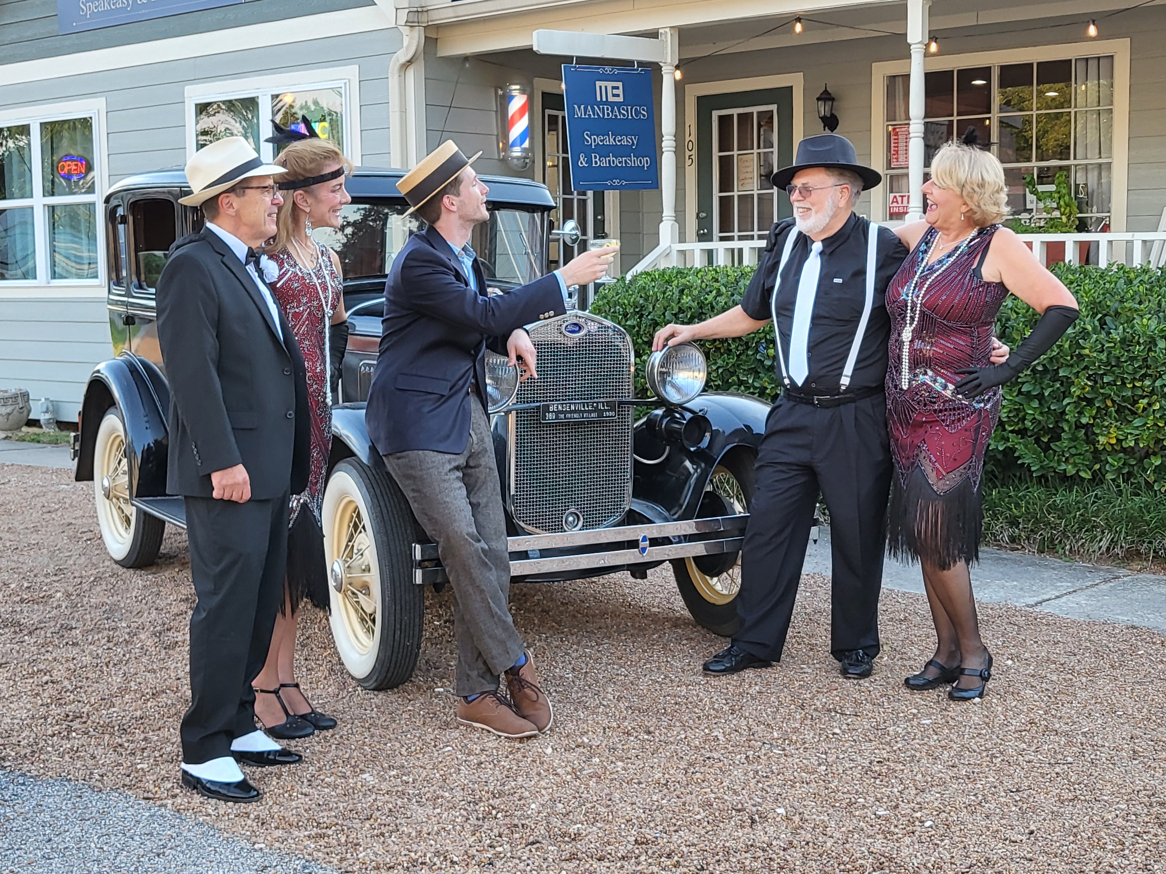 Five people in Roaring Twenties outfits — men in fedoras, suspenders and bow ties and women in beaded flapper dresses — laughing and posing around a black vintage 1920s car parked outside a speakeasy-style storefront on a small-town main street.