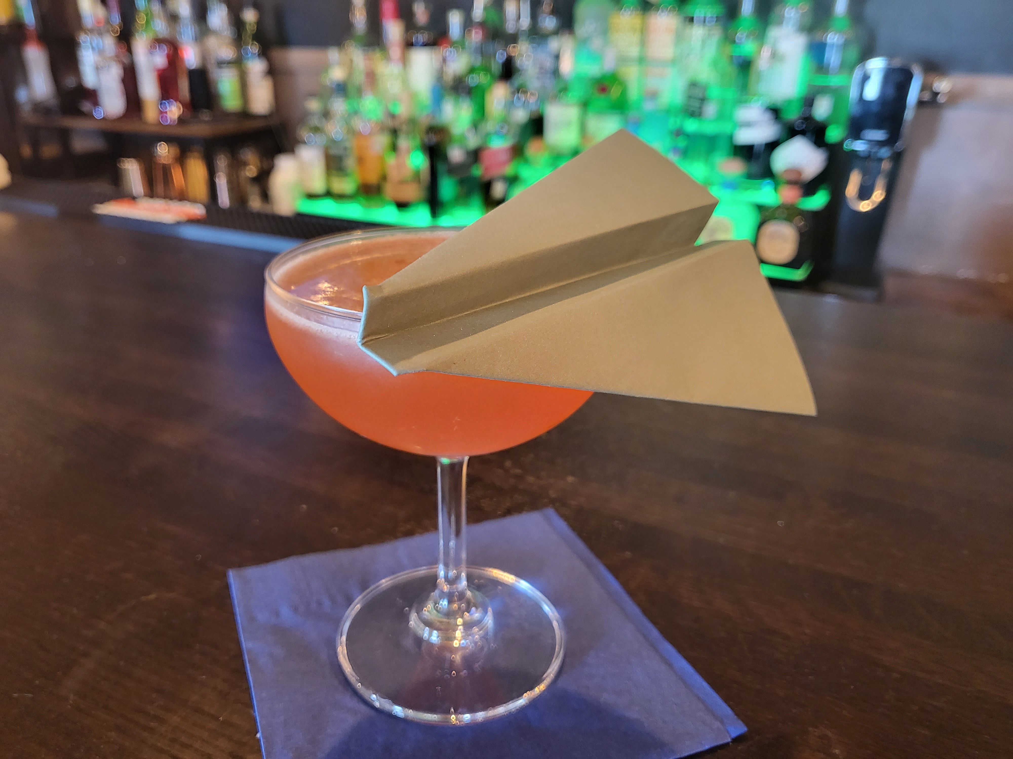 Playful paper-airplane garnish perched on a pink cocktail in a coupe glass, resting on a blue napkin on a wooden bar counter with blurred bottles in the background.