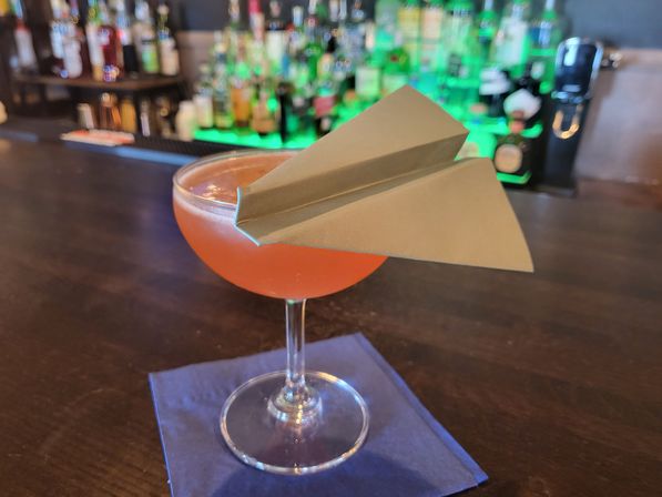 Playful paper-airplane garnish perched on a pink cocktail in a coupe glass, resting on a blue napkin on a wooden bar counter with blurred bottles in the background.