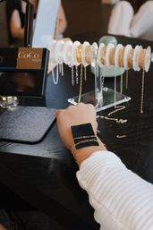 Wrist wearing delicate gold chain bracelets being tested against a display rack of gold and silver chains on a dark wood boutique table.
